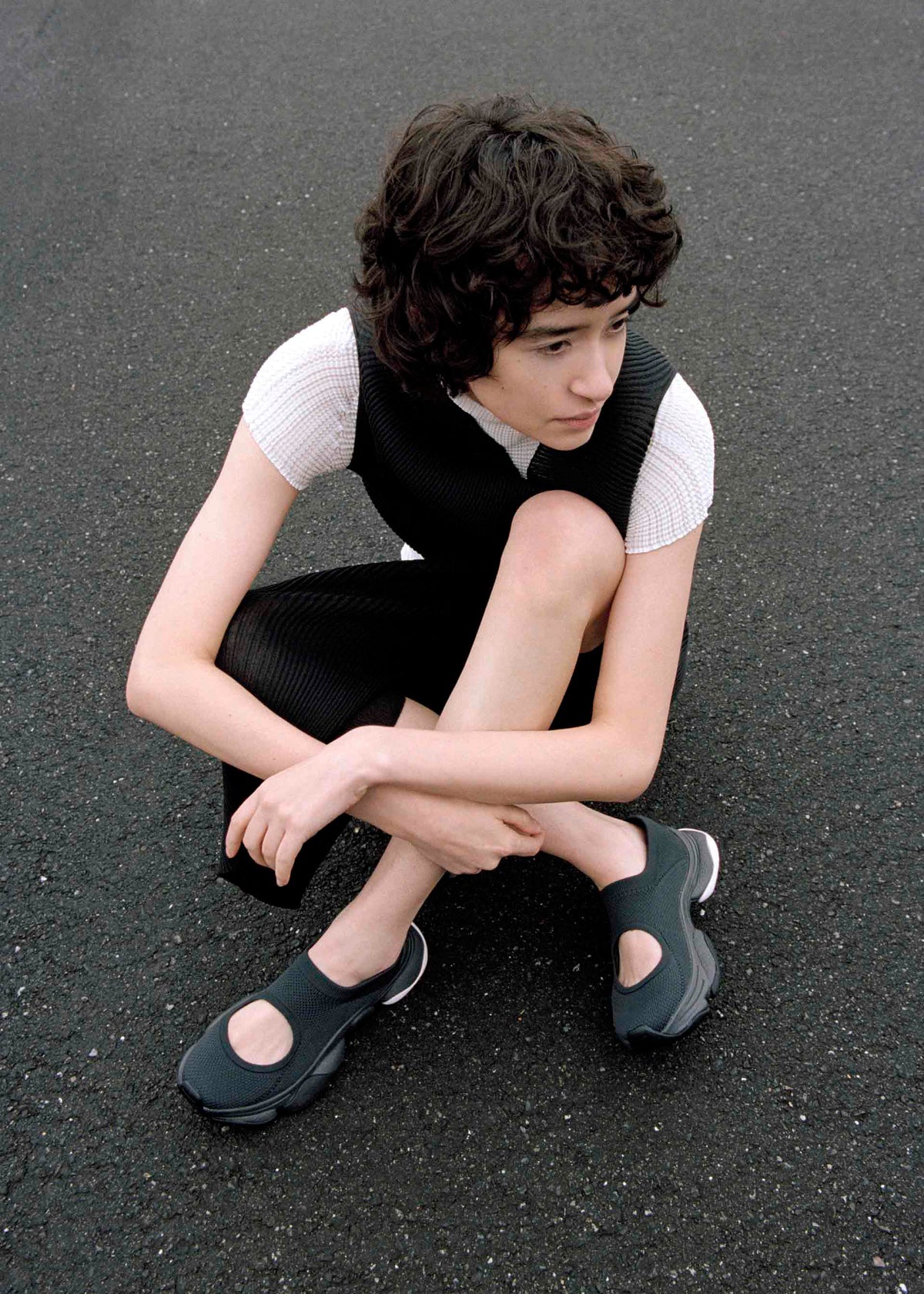 A person with short curly hair sits on asphalt, wearing ISSEY MIYAKE's KARST FINCH black sleeveless dress over a white top and colorful sneakers with cutouts, looking to the side with one arm resting on their knee. view 16 of 18