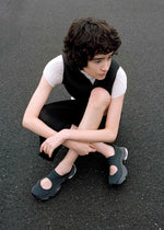 A person with short curly hair sits on asphalt, wearing a short-sleeve white top under a black dress and black ISSEY MIYAKE KARST FINCH sneakers with cutout details, resting one arm on their knee and looking to the side.