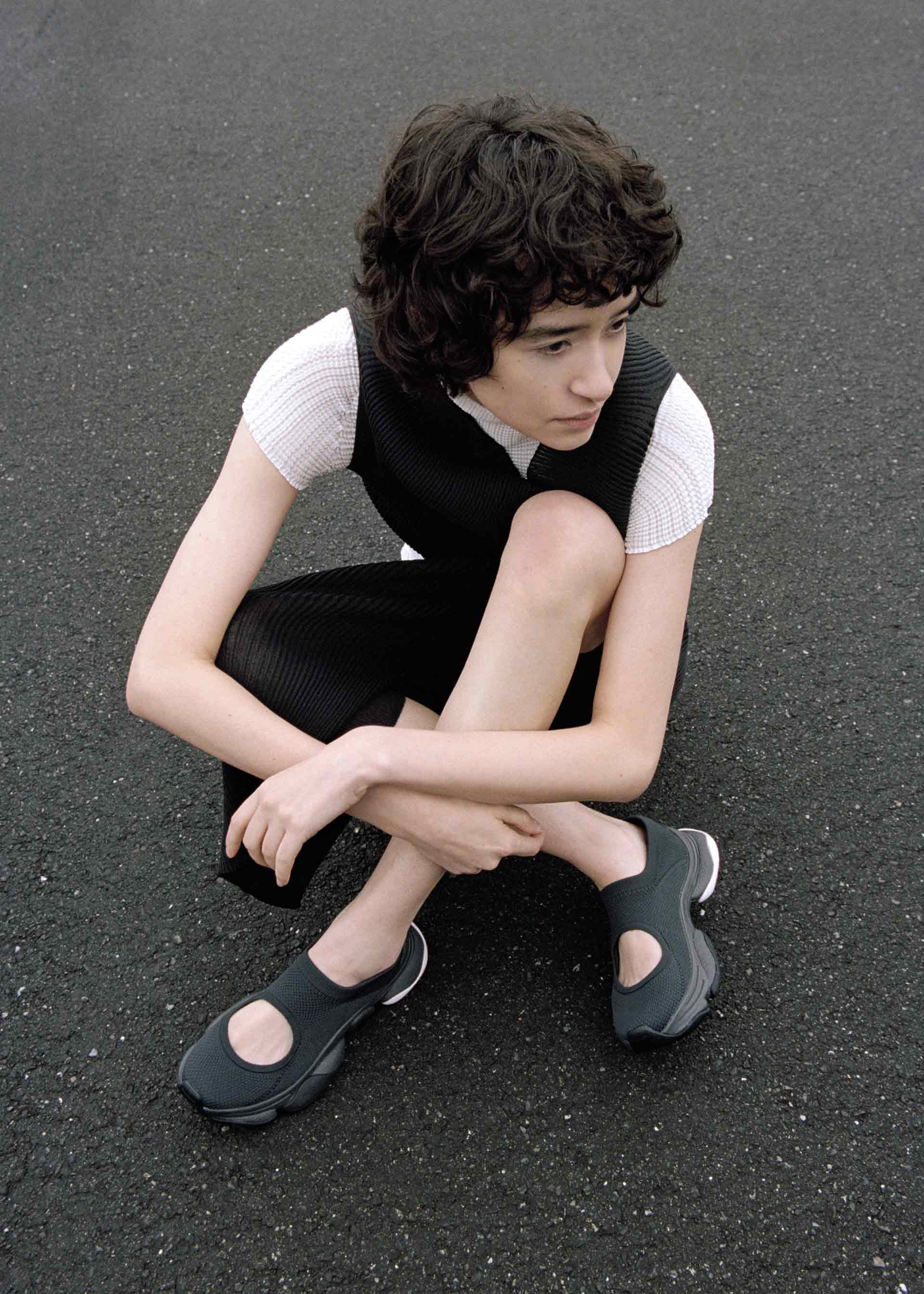 Zoomed view A person with short curly hair sits on asphalt, wearing a short-sleeve white top under a black dress and black ISSEY MIYAKE KARST FINCH sneakers with cutout details, resting one arm on their knee and looking to the side.
