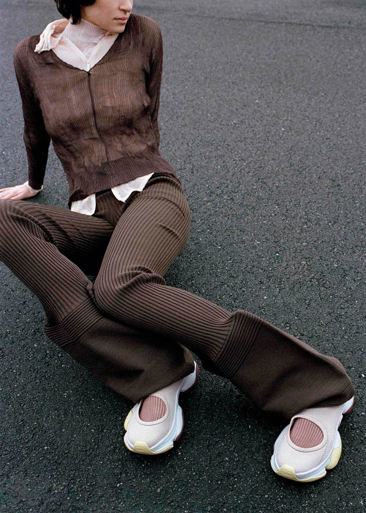 A person in a brown ribbed jacket and flared pants sits on asphalt, legs outstretched, wearing oversized ISSEY MIYAKE KARST FINCH sneakers in white and pink. Their face is partly turned away, with a sheer scarf around their neck. view 18 of 18