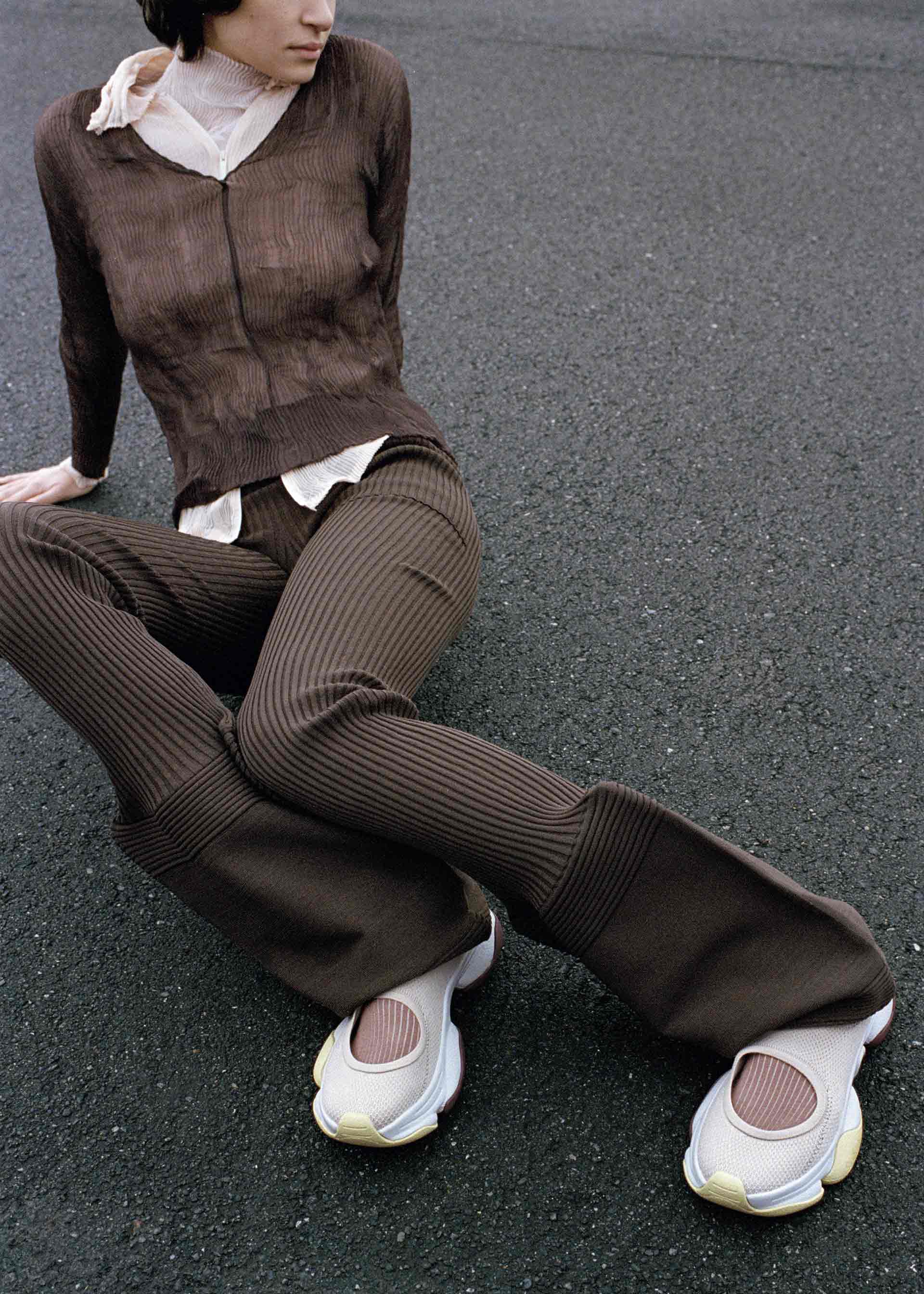 Zoomed view A person in a brown ribbed jacket and flared pants sits on asphalt, legs outstretched, wearing oversized ISSEY MIYAKE KARST FINCH sneakers in white and pink. Their face is partly turned away, with a sheer scarf around their neck.