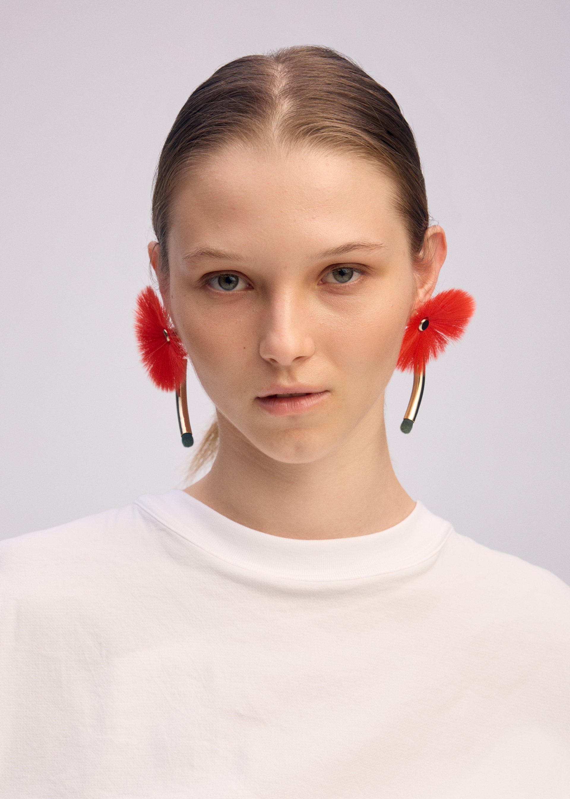 Zoomed view A person with straight, light brown hair pulled back wears a plain white top and large, bright red ENGINEERED FLOWER ACC earrings by ISSEY MIYAKE. The background is a solid light color.