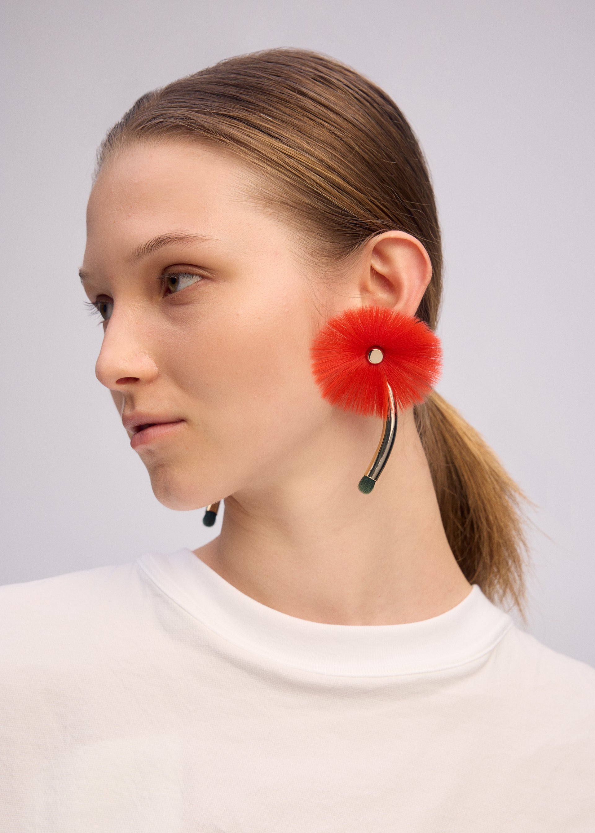 Zoomed view A person with light skin and straight brown hair tied back wears the ISSEY MIYAKE ENGINEERED FLOWER ACC, a handcrafted earring inspired by traditional Japanese jewelry, shaped like a fluffy red flower with a curved stem and green tip. They wear a plain white top.