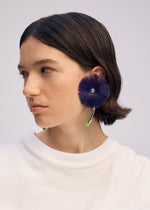 A woman with short brown hair wears a white shirt and ISSEY MIYAKE ENGINEERED FLOWER ACC: large, round, fluffy dark purple earrings with a silver center and green tip, inspired by Kumano brush designs, against a plain light background.