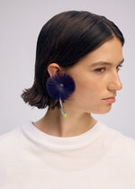 A person with short brown hair wears a white shirt and a large, round ISSEY MIYAKE ENGINEERED FLOWER ACC earring with a curved metallic accent and green tip, inspired by traditional Japanese jewelry.
