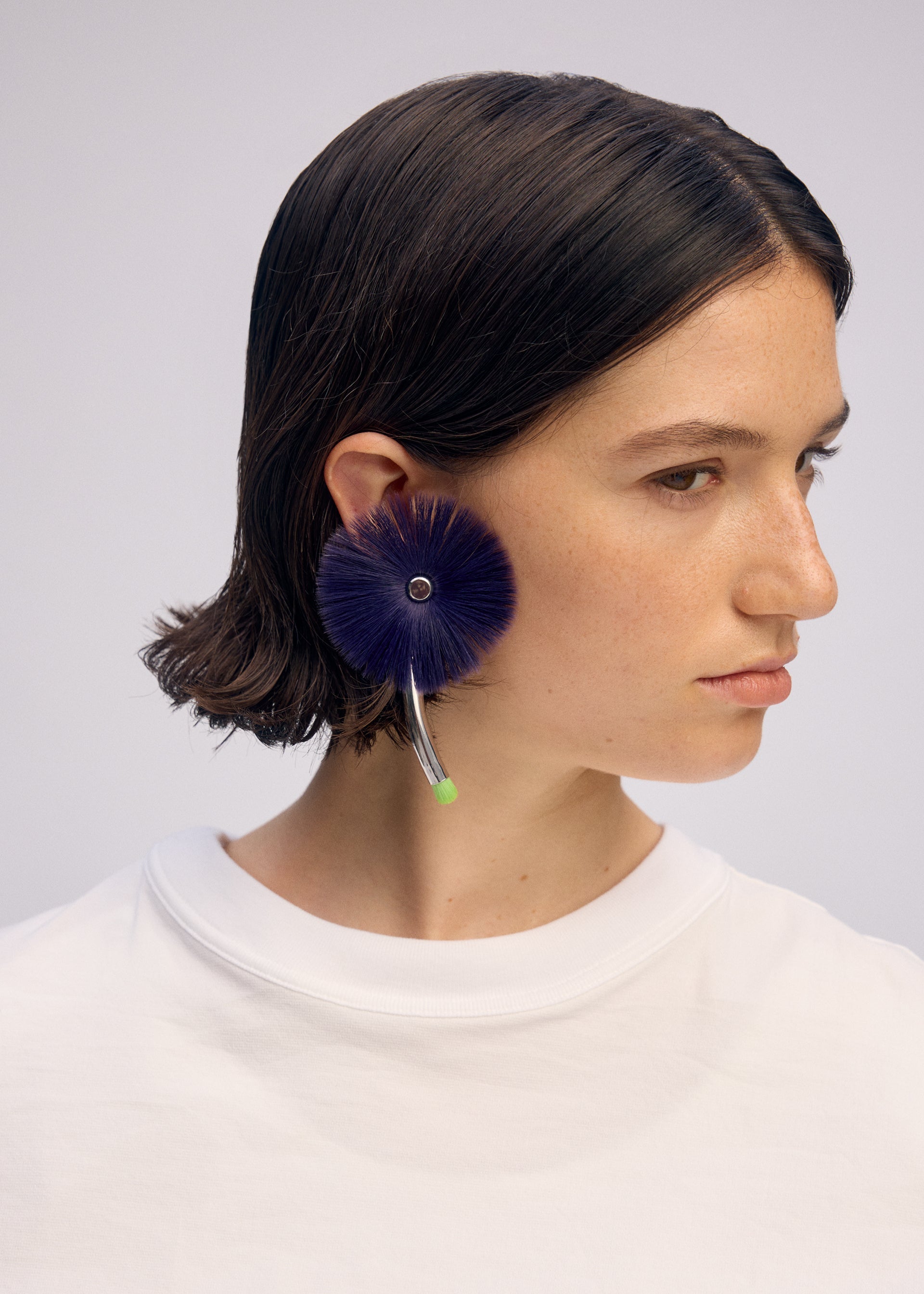 Zoomed view A person with short brown hair wears a white shirt and a large, round ISSEY MIYAKE ENGINEERED FLOWER ACC earring with a curved metallic accent and green tip, inspired by traditional Japanese jewelry.