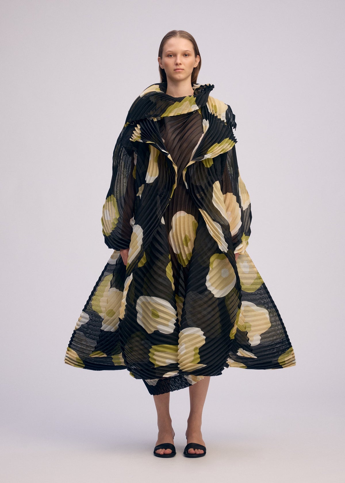 A person stands facing forward, wearing the ISSEY MIYAKE VERVE coat, which features handmade pleats, bold yellow and white hand-drawn floral patterns, voluminous sleeves, a flared silhouette, black sandals, and straight loose hair. view 5 of 9