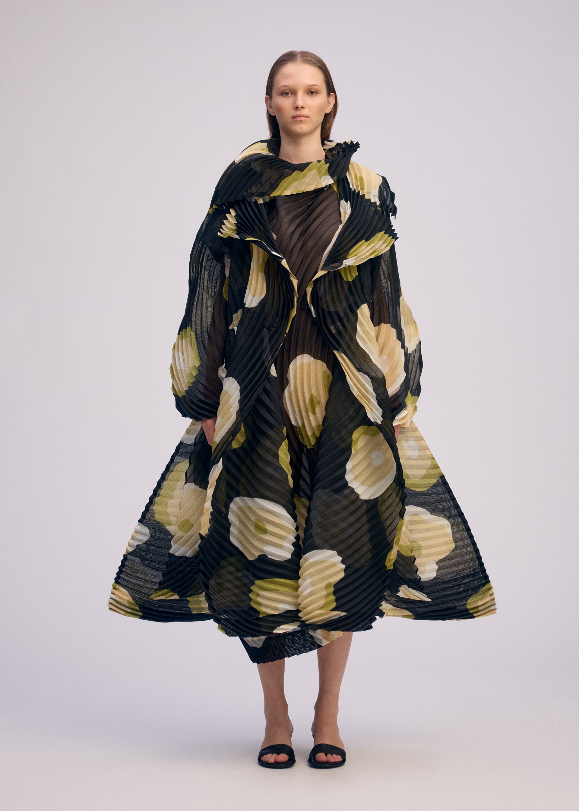 Zoomed view A person stands facing forward, wearing the ISSEY MIYAKE VERVE coat, which features handmade pleats, bold yellow and white hand-drawn floral patterns, voluminous sleeves, a flared silhouette, black sandals, and straight loose hair.