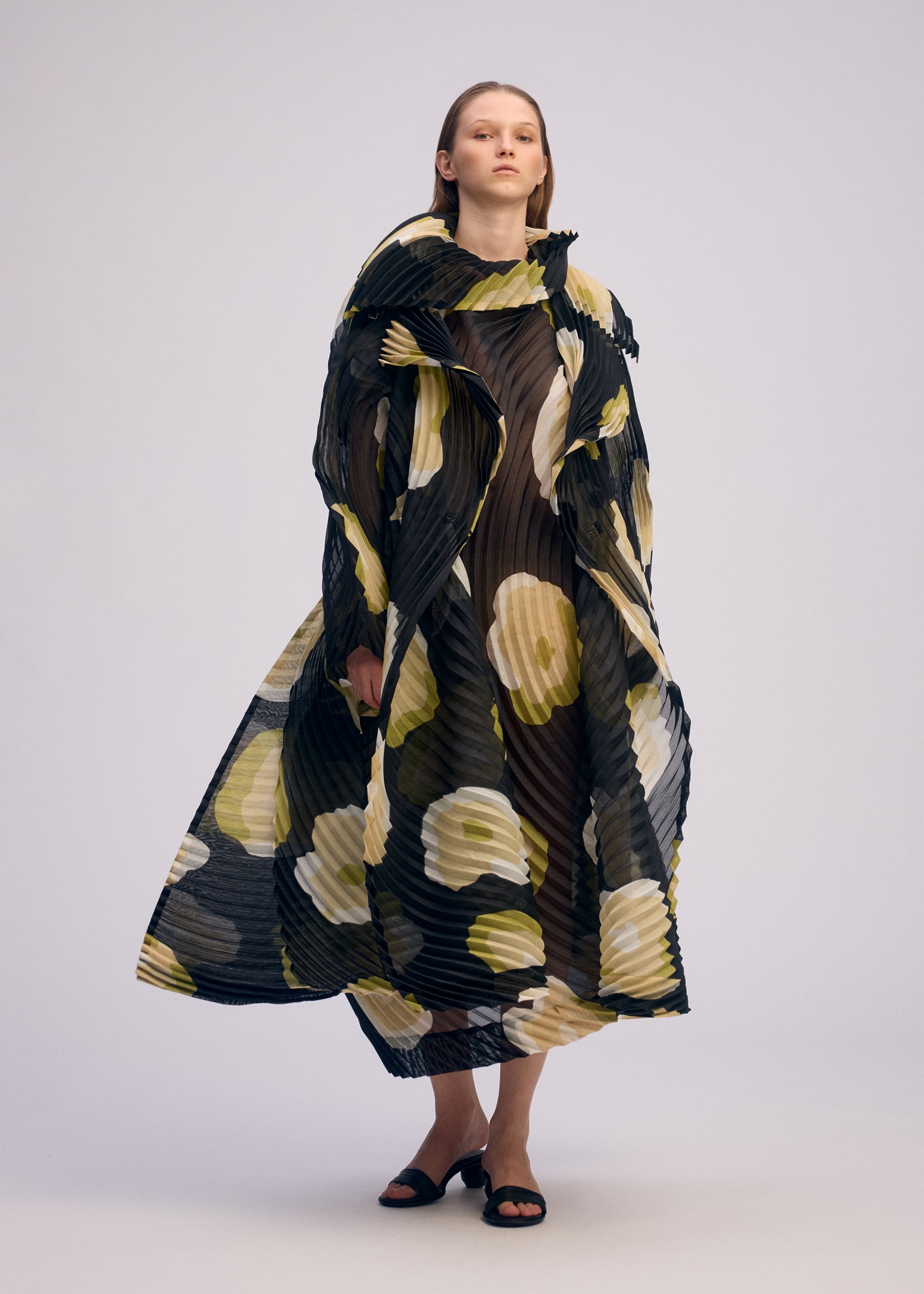 Zoomed view A person stands against a plain background wearing the VERVE dress by ISSEY MIYAKE—a sheer, pleated black dress with handmade pleats and bold yellow and white hand-drawn floral patterns, layered over a black underdress and paired with black sandals.