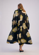 A person with straight, light brown hair models the ISSEY MIYAKE VERVE coat, a long pleated piece with bold white and green floral patterns, standing against a plain light background and facing away from the camera.