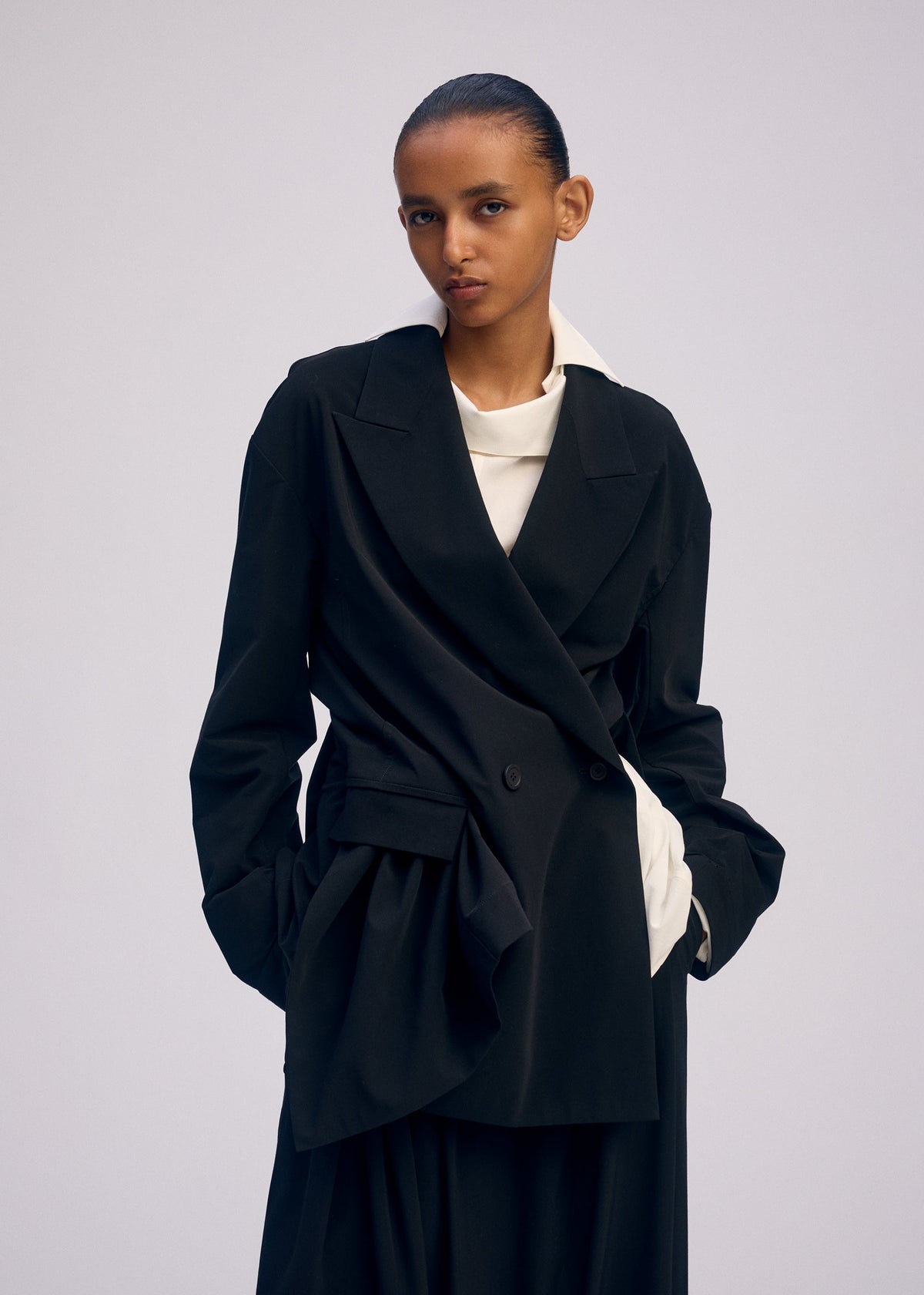 A person with slicked-back hair wears the ISSEY MIYAKE LOOPED loose-fitting, hand washable black double-breasted blazer over a white top, standing with hands in pockets against a plain, light background. view 5 of 5
