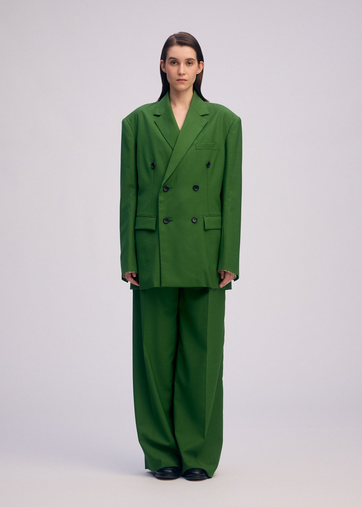 A person with straight brown hair faces forward, wearing an oversized green ISSEY MIYAKE PALINDROME jacket and black shoes, standing against a plain light grey background. view 8 of 15