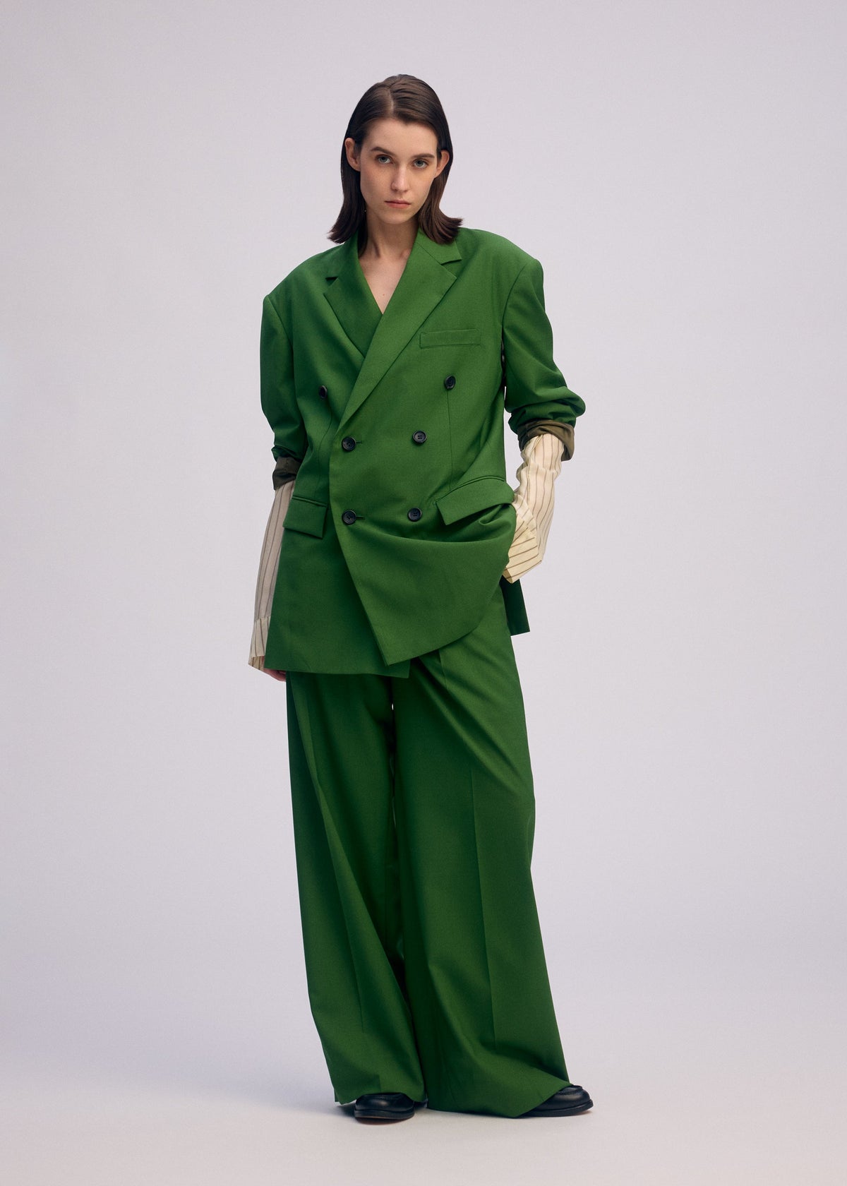 A person stands against a plain background wearing the oversized green ISSEY MIYAKE PALINDROME jacket, styled with wide-leg pants and a double-breasted blazer over a cream ribbed top, hands in pockets and a neutral expression. view 9 of 15