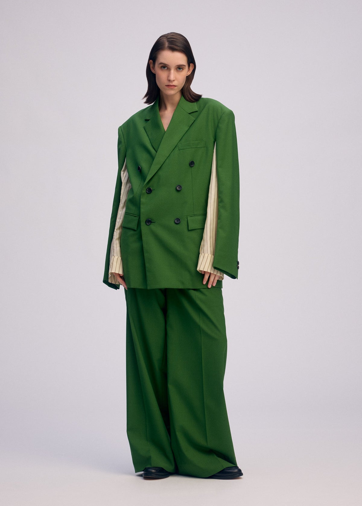 A person stands against a plain background wearing ISSEY MIYAKE’s PALINDROME jacket with pinstriped inner sleeves, wide-legged pants, and black shoes, looking directly at the camera with a neutral expression. view 10 of 15