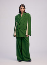 A person stands against a plain background wearing ISSEY MIYAKE’s PALINDROME jacket with pinstriped inner sleeves, wide-legged pants, and black shoes, looking directly at the camera with a neutral expression.