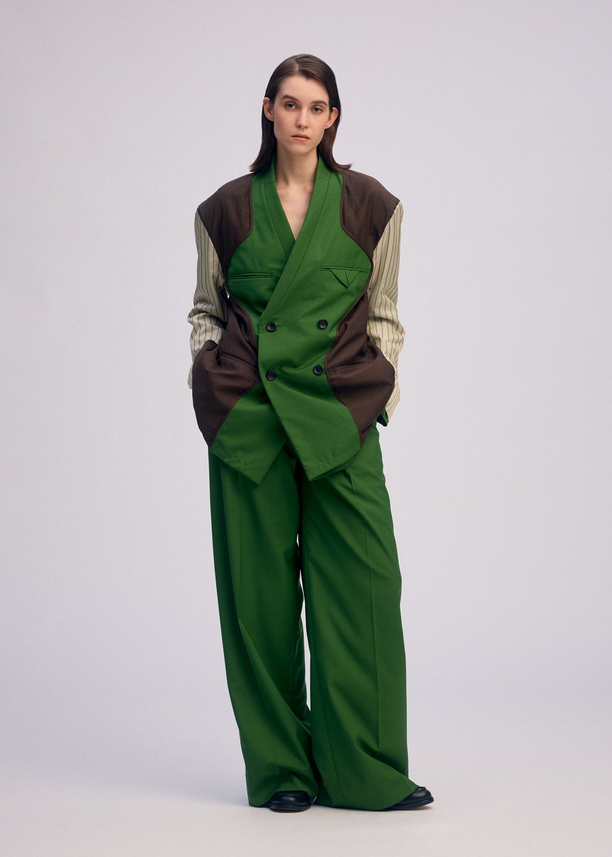 A person with straight, dark hair wears a wide-legged green suit and the ISSEY MIYAKE PALINDROME jacket with brown panels and striped sleeves, standing hands-in-pockets against a plain light background. view 11 of 15