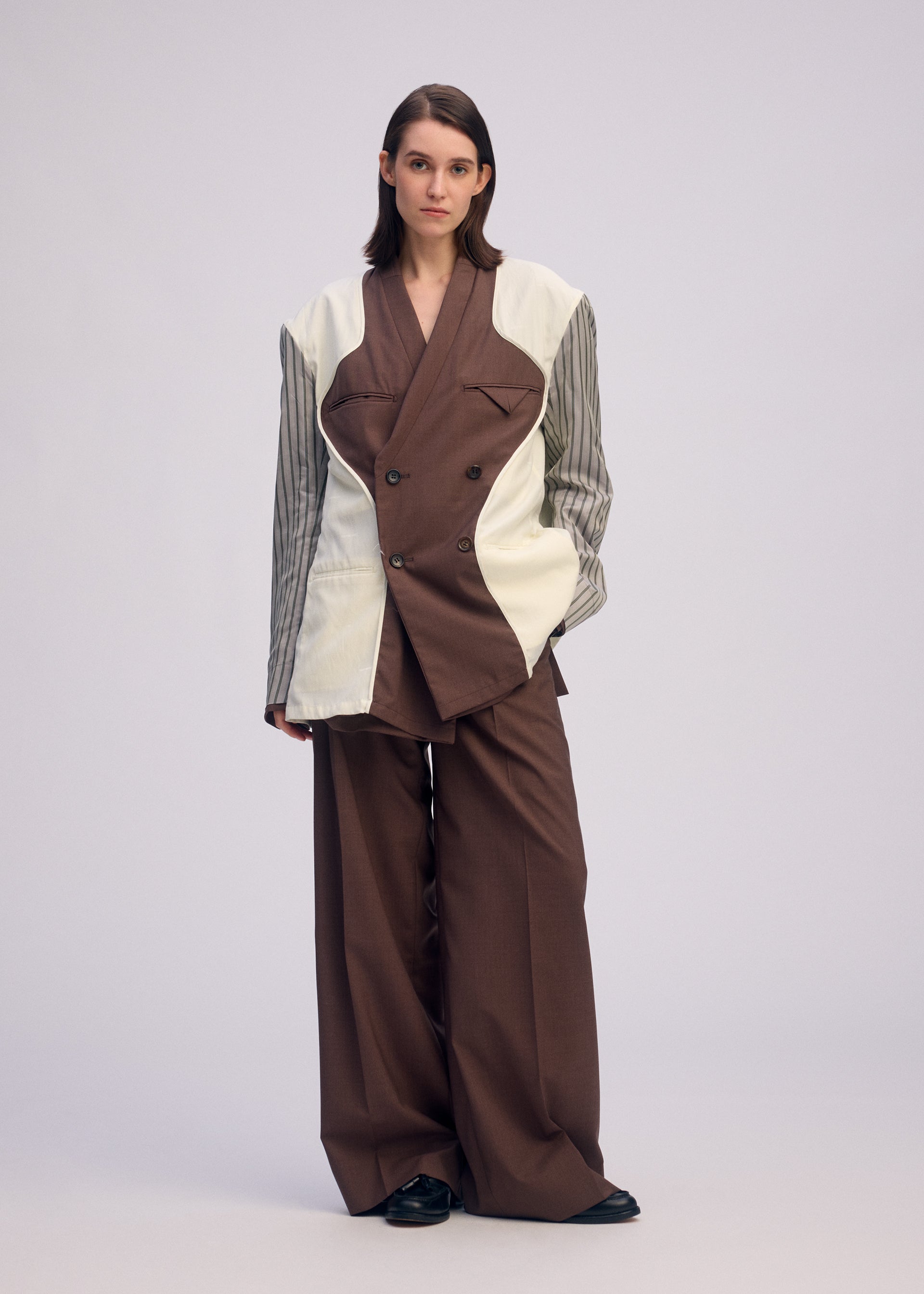 Zoomed view A person with straight brown hair and a neutral expression wears ISSEY MIYAKE's PALINDROME: an oversized brown suit with wide-legged pants and a unique jacket featuring cream and striped panels, standing against a light background.