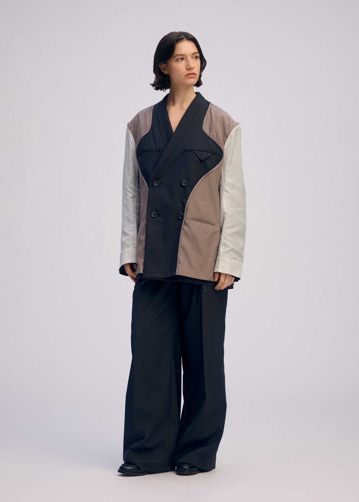 A person with medium-length dark hair wears the ISSEY MIYAKE PALINDROME jacket in black, gray, and beige, styled with oversized black trousers and shoes, standing against a plain light background. view 13 of 15