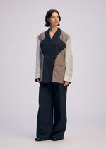 A person with medium-length dark hair wears the ISSEY MIYAKE PALINDROME jacket in black, gray, and beige, styled with oversized black trousers and shoes, standing against a plain light background.
