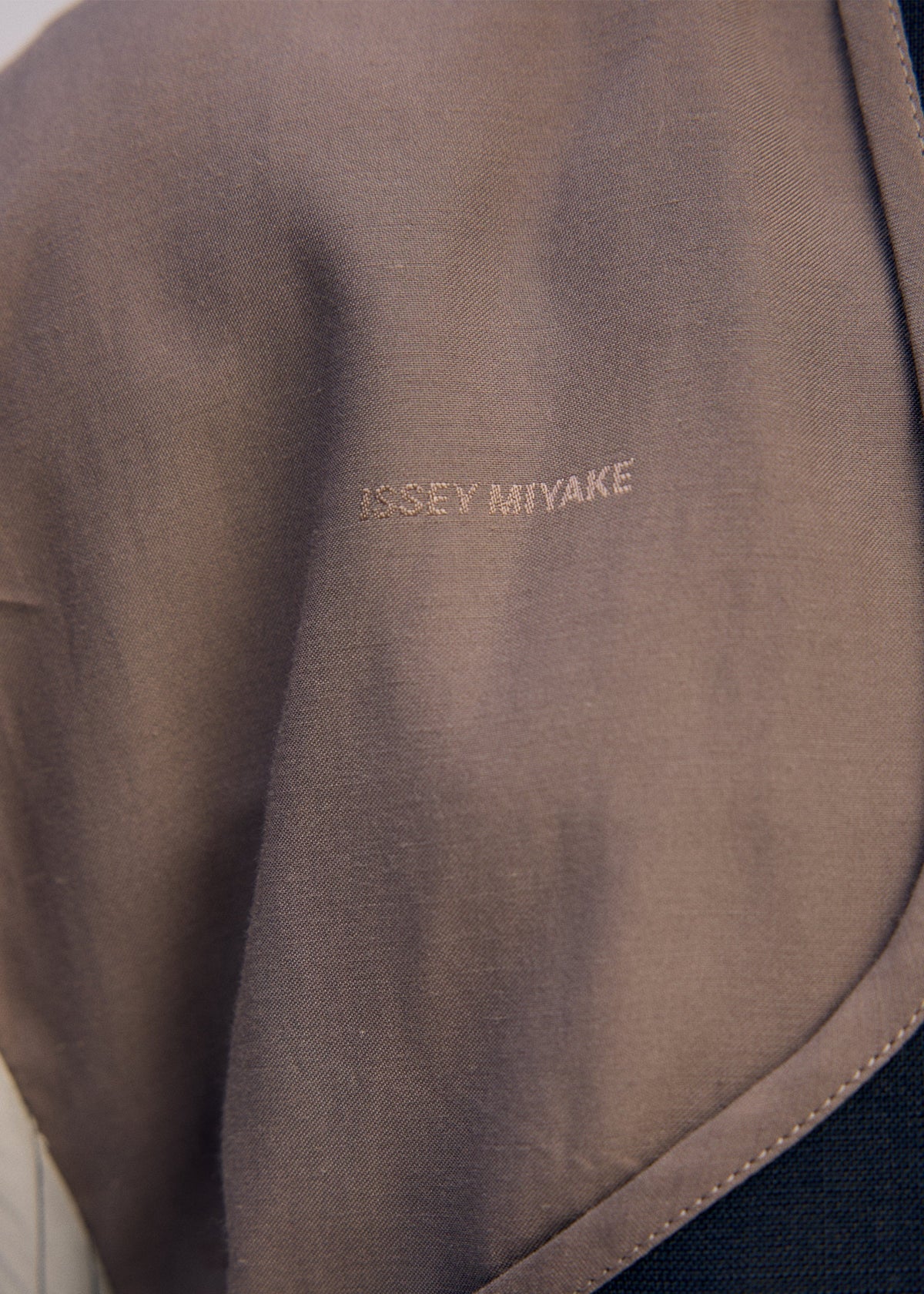 A close-up of a fabric label displays "ISSEY MIYAKE" embroidered in light thread on smooth, taupe cloth, showcasing the craftsmanship of the ISSEY MIYAKE PALINDROME jacket. view 14 of 15