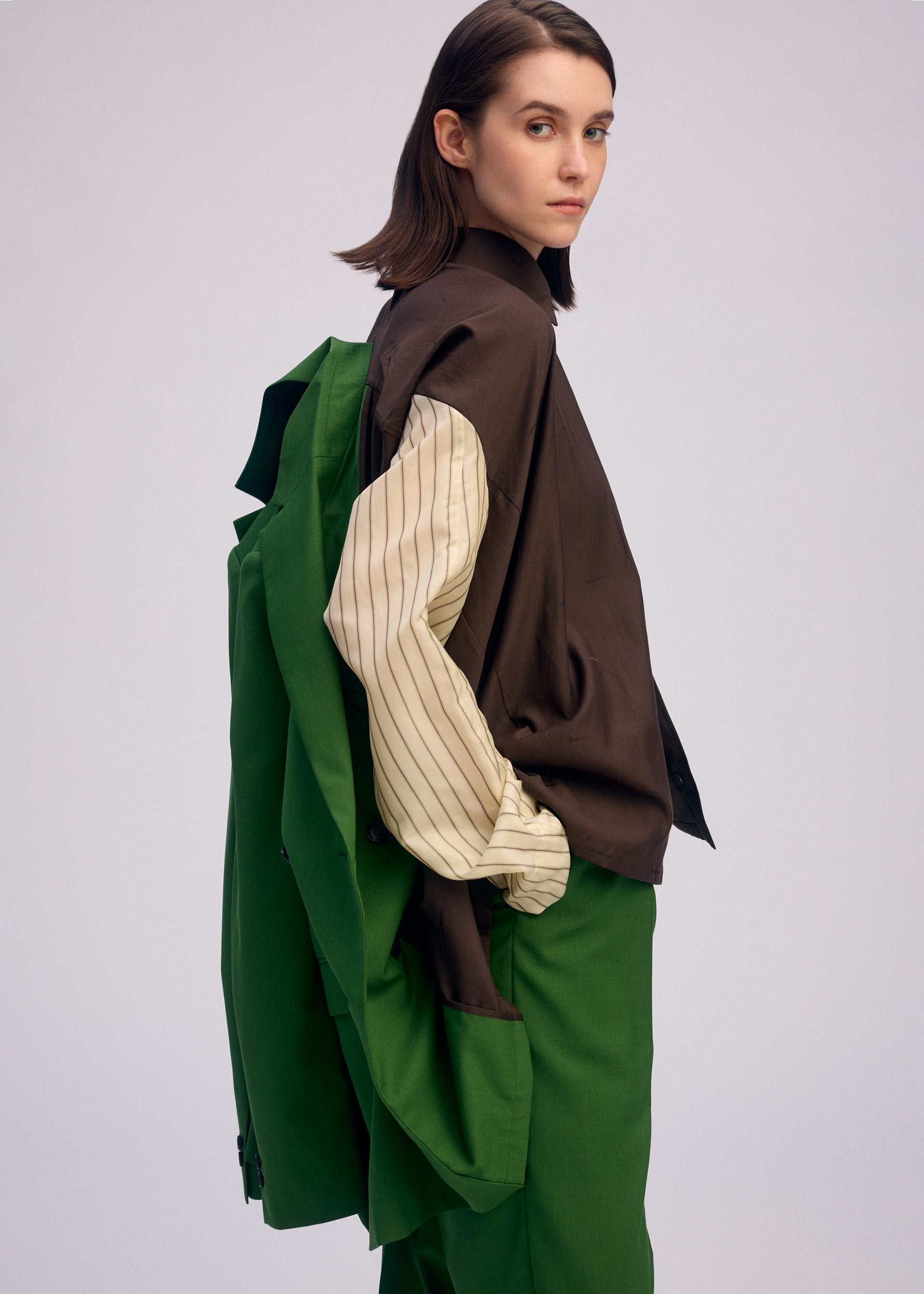 Zoomed view A woman with straight brown hair wears the ISSEY MIYAKE PALINDROME jacket draped over her shoulder, paired with green pants and layered shirts. She looks sideways with a neutral expression against a plain background.