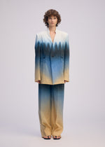 A person models ISSEY MIYAKE's PLUMAGE: a collarless suit with a five-color gradient from cream to blue and black, evoking hand-painted Kyoto artistry, paired with open-toed black sandals against a plain background.