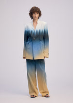 A person with curly hair wears the ISSEY MIYAKE PLUMAGE collarless jacket and loose suit in a five-color gradient, buttoned to reveal a deep V-neck, standing against a plain light background.