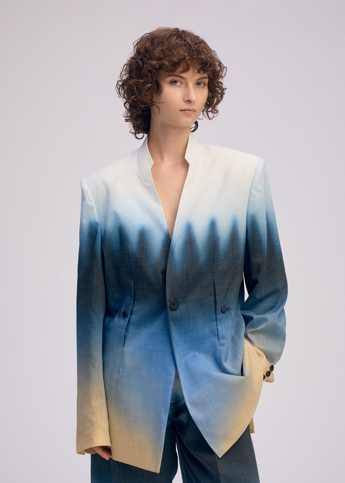 A person with curly brown hair models ISSEY MIYAKE’s PLUMAGE: a loose-fitting, five-color gradient collarless jacket and matching pants, posing with one hand in their pocket against a plain light background—Kyoto-inspired elegance. view 6 of 6