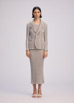 A woman with short, dark hair and a neutral expression wears the ISSEY MIYAKE WAVELET ENSEMBLE— a tailored jacquard stretch jacket and long skirt—paired with white open-toe sandals, standing against a plain background.