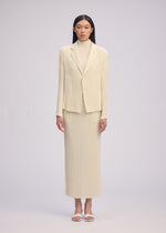 A woman faces forward in a cream HATCHING PLEATS jacket over a matching turtleneck and ankle-length skirt by ISSEY MIYAKE, styled with straight dark hair and white sandals against a plain light background.