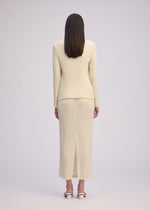 A person with straight brown hair faces away, wearing the ISSEY MIYAKE HATCHING PLEATS jacket and cream textured skirt suit made from recycled polyester, paired with open-toed sandals against a plain light background.