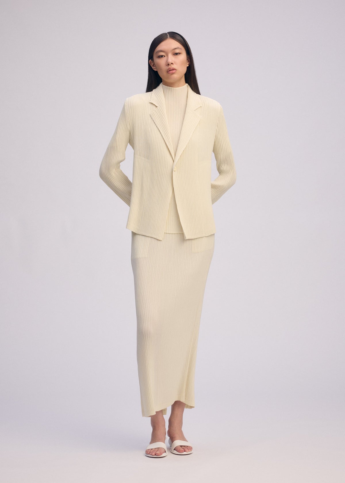 A woman stands facing forward in ISSEY MIYAKE's HATCHING PLEATS: a cream ribbed suit with pleated jacket, matching long skirt, and turtleneck. She wears white open-toe heels; her long hair is straight against a plain background. view 7 of 8