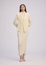 A woman stands facing forward in ISSEY MIYAKE's HATCHING PLEATS: a cream ribbed suit with pleated jacket, matching long skirt, and turtleneck. She wears white open-toe heels; her long hair is straight against a plain background.