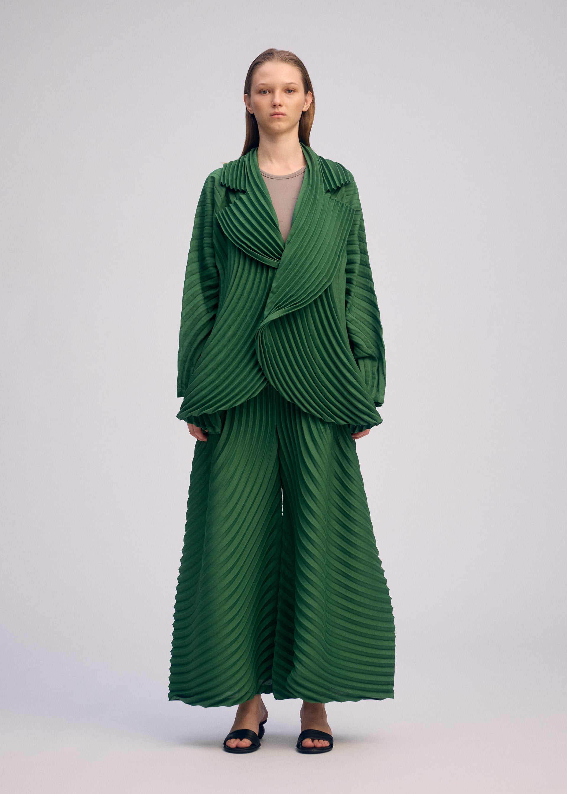 Zoomed view A person poses against a plain background, wearing the ISSEY MIYAKE CIRCLE PLEATS PB set in green with handmade pleats, black sandals, and straight hair worn down.