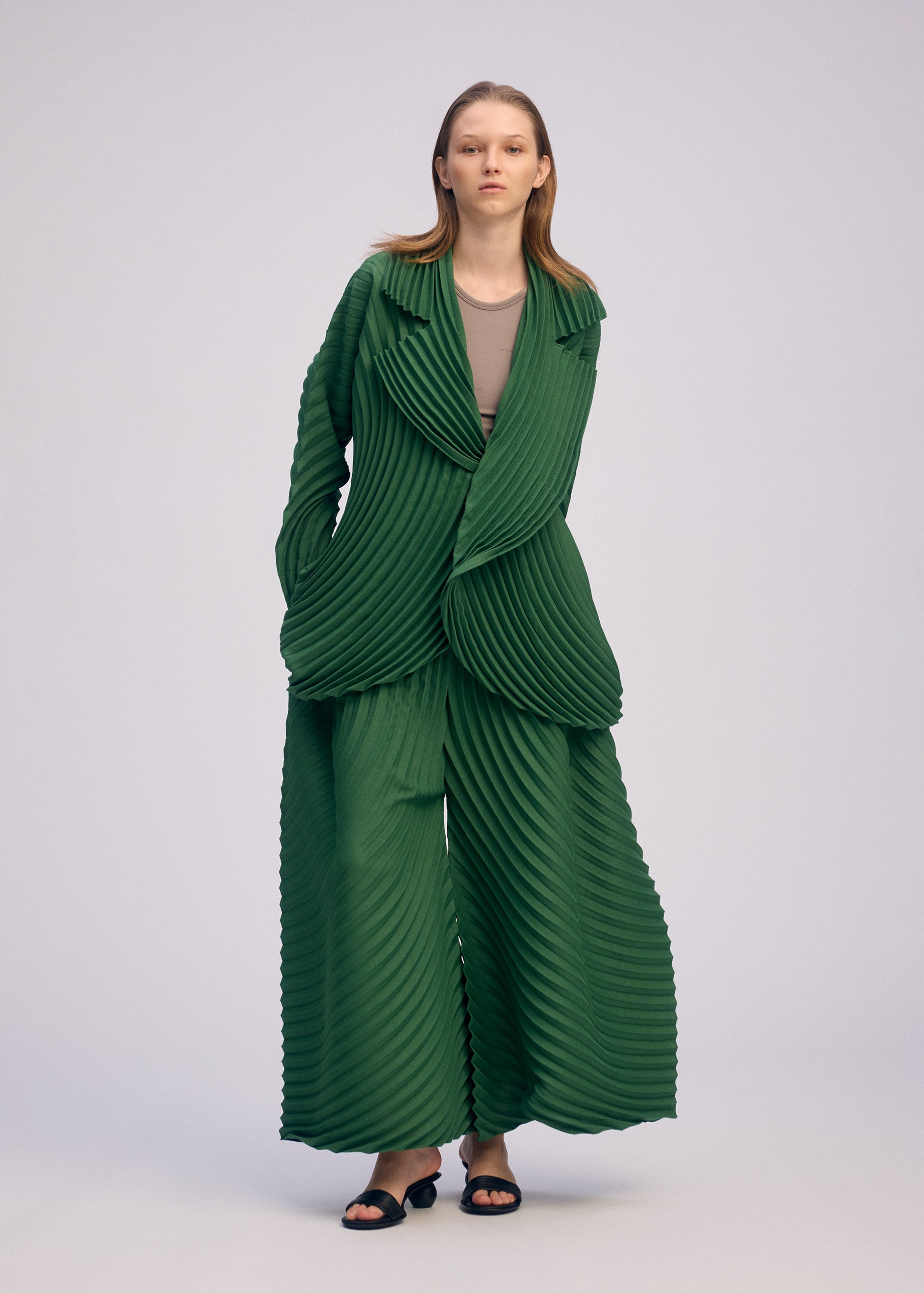 Zoomed view A person stands against a plain background wearing ISSEY MIYAKE's CIRCLE PLEATS PB: an oversized, handmade pleated green suit with wide-legged pants and matching jacket over a taupe shirt, paired with black sandals.