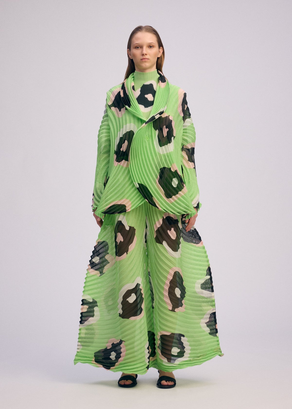 A person wears ISSEY MIYAKE's VERVE: a loose, pleated set in bright green with bold black, white, and brown spots. The wide-leg pants and draped high-neck top showcase handmade pleats. The background is plain and light-colored. view 5 of 9
