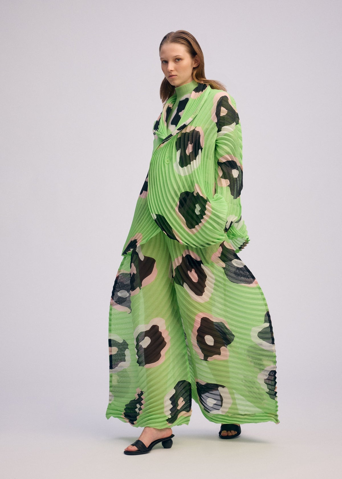 A woman showcases the ISSEY MIYAKE VERVE set—a loose, pleated ensemble with wide-leg pants. The long-sleeved top displays bold black, beige, and pink abstract spots on a light base, paired with black heeled sandals. view 6 of 9