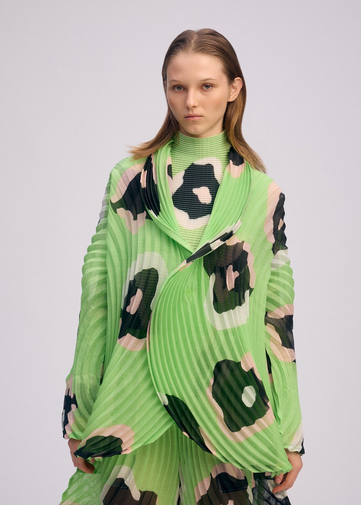 A person with straight, shoulder-length hair wears the ISSEY MIYAKE VERVE jacket in bright green with bold black and beige hand-drawn floral pleats, standing against a plain light background. view 7 of 9