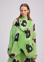 A person with straight, shoulder-length hair wears the ISSEY MIYAKE VERVE jacket in bright green with bold black and beige hand-drawn floral pleats, standing against a plain light background.