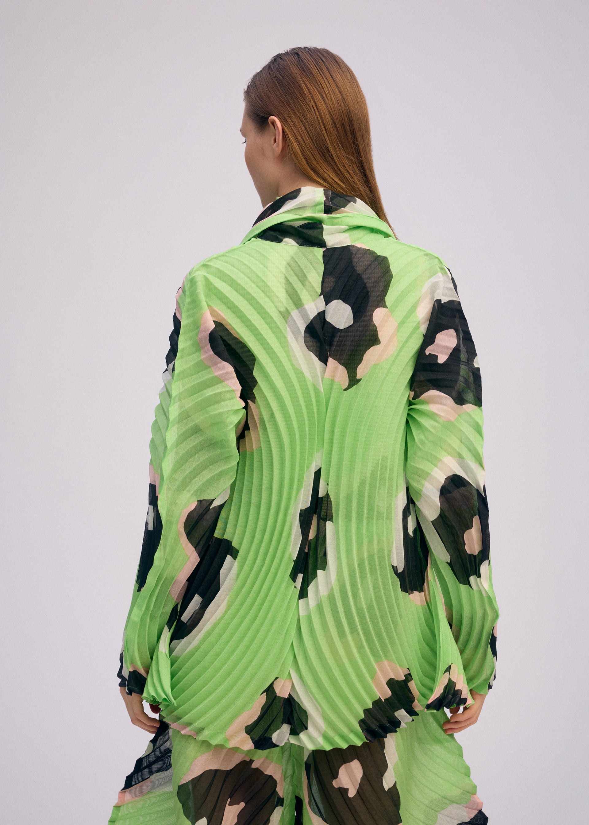 Zoomed view A person with long, straight brown hair is seen from behind wearing the VERVE pleated jacket and matching floral pants by ISSEY MIYAKE in green, black, and beige against a plain light background.