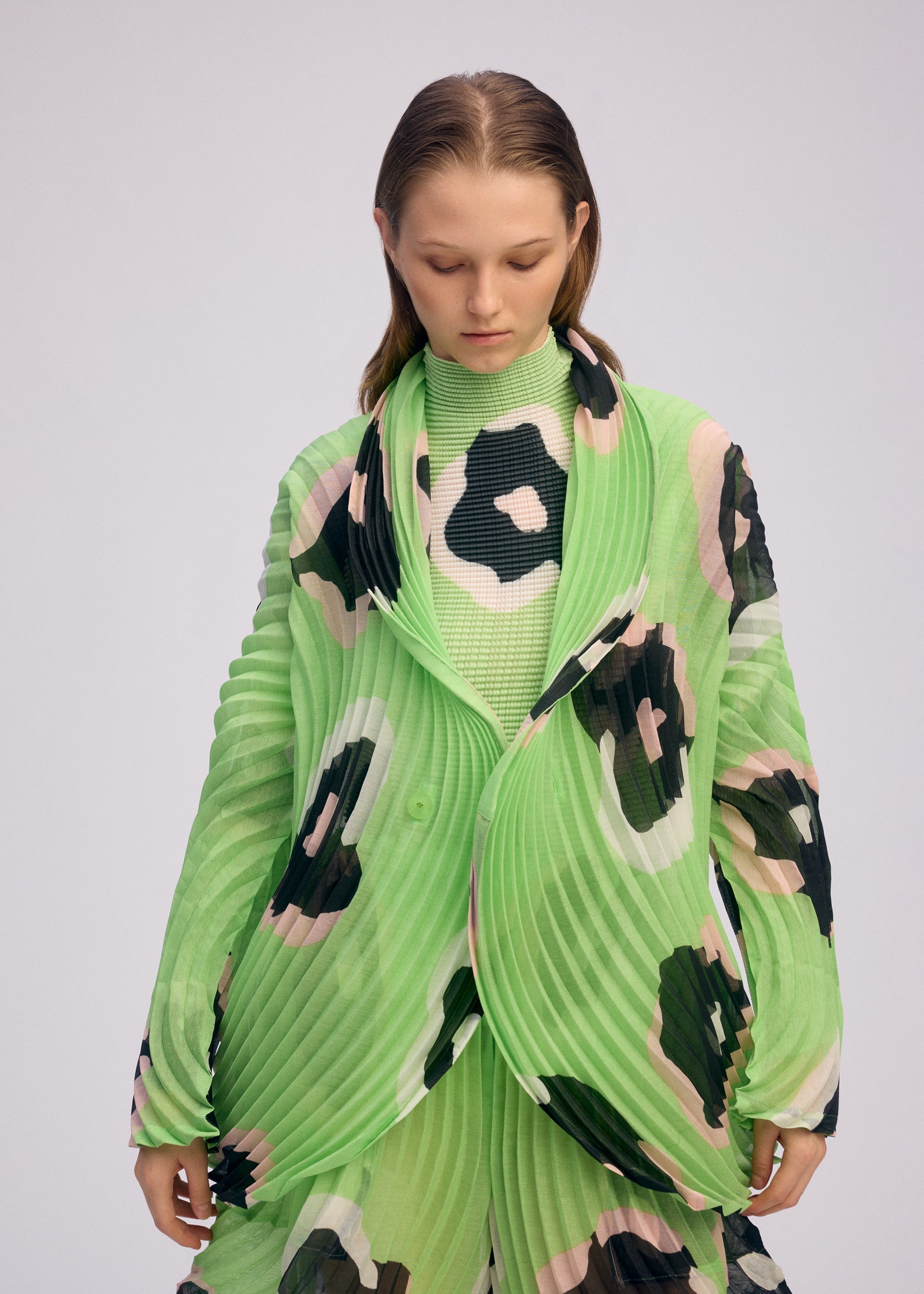 Zoomed view A person with straight, light brown hair looks down while wearing the VERVE pleated jacket by ISSEY MIYAKE in bright green with bold black, white, and pink hand-drawn floral patterns. The background is plain and light-colored.