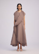 A woman with straight dark hair wears ISSEY MIYAKE’s CIRCLE PLEATS PB taupe, textured dress with handmade pleats, asymmetrical layers, draped scarf detail, and long vest. She pairs white sandals against a plain light background.