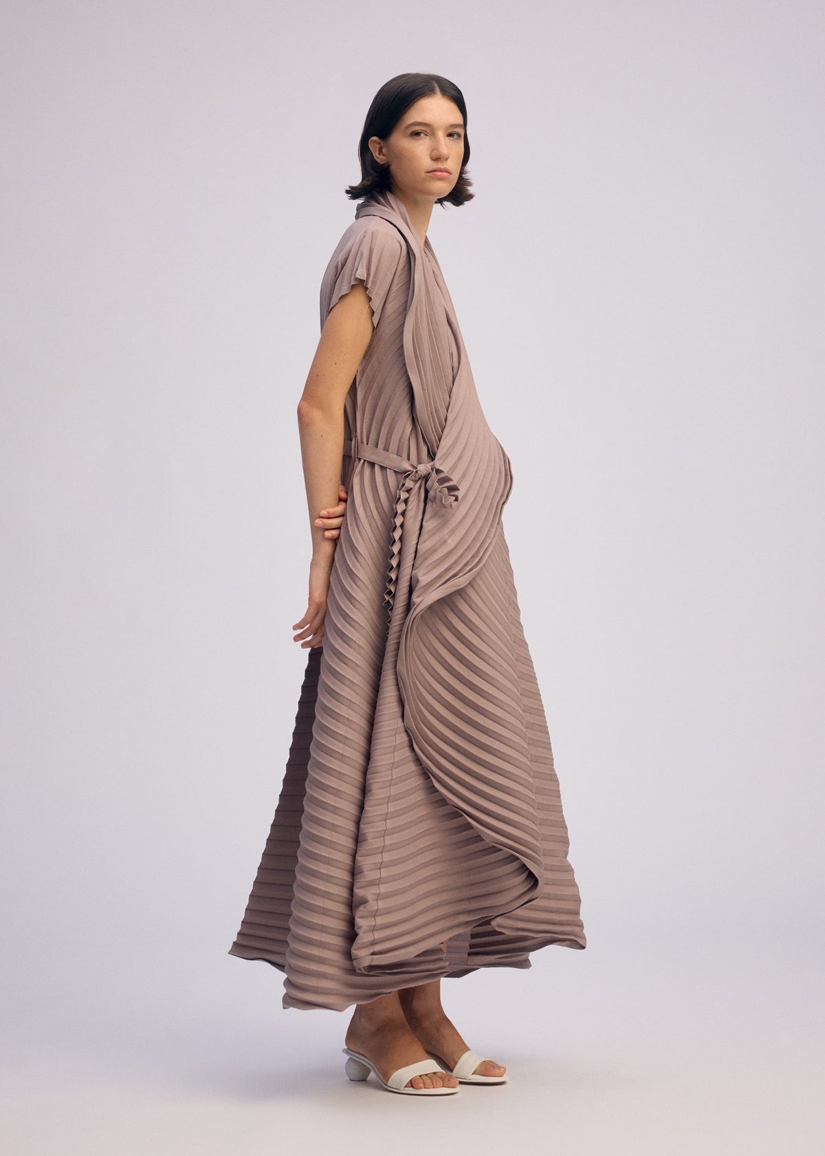A woman with short dark hair wears the ISSEY MIYAKE CIRCLE PLEATS PB dress in mauve, featuring handmade pleats, a wrap design, and cap sleeves. She stands sideways in white sandals and a minimal bracelet against a light background. view 6 of 6