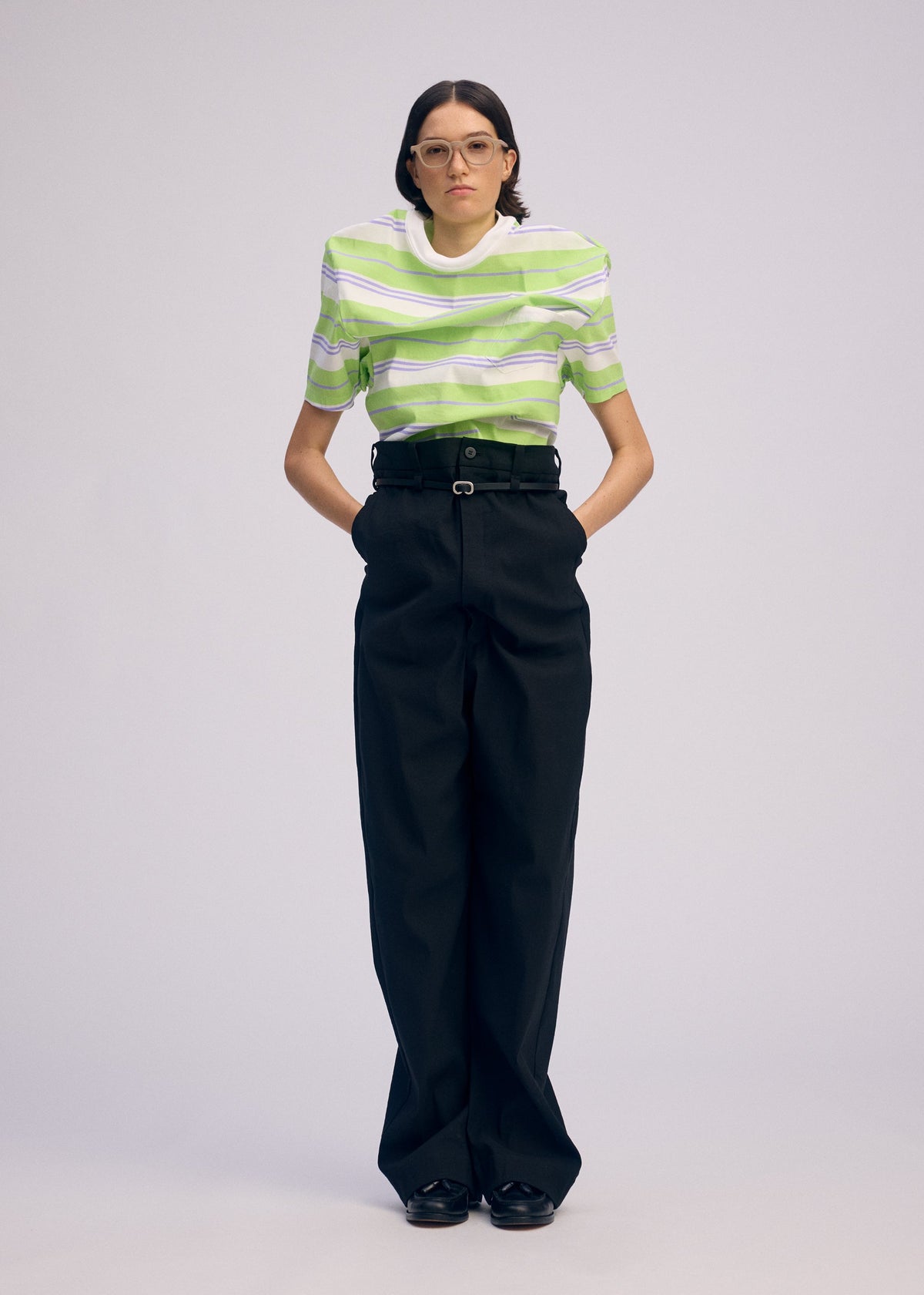 A person stands against a plain background wearing glasses, a green and white striped short-sleeve shirt, ISSEY MIYAKE GENERIC WEAR high-waisted black wide-leg trousers, black shoes, and has their hands behind their back. view 4 of 7