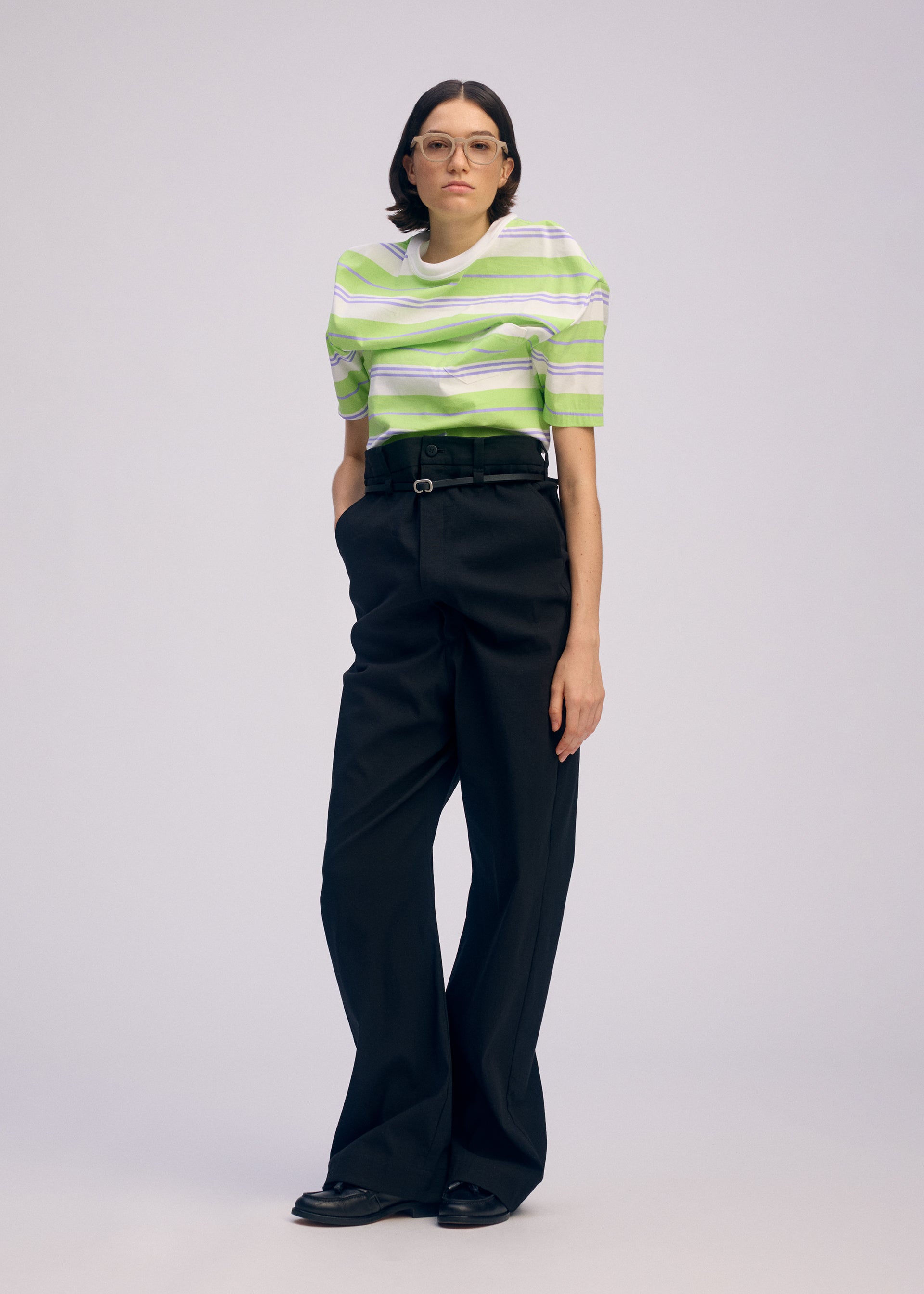 Zoomed view A person models GENERIC WEAR by ISSEY MIYAKE: glasses, a green and white striped short-sleeve shirt, and playful high-waisted pants, standing with hands in pockets against a plain light background.