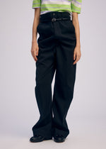 Wearing ISSEY MIYAKE GENERIC WEAR: a green and white striped short-sleeve shirt, loose high-waisted black trousers with a black belt, and black shoes against a plain light background. Head not visible.