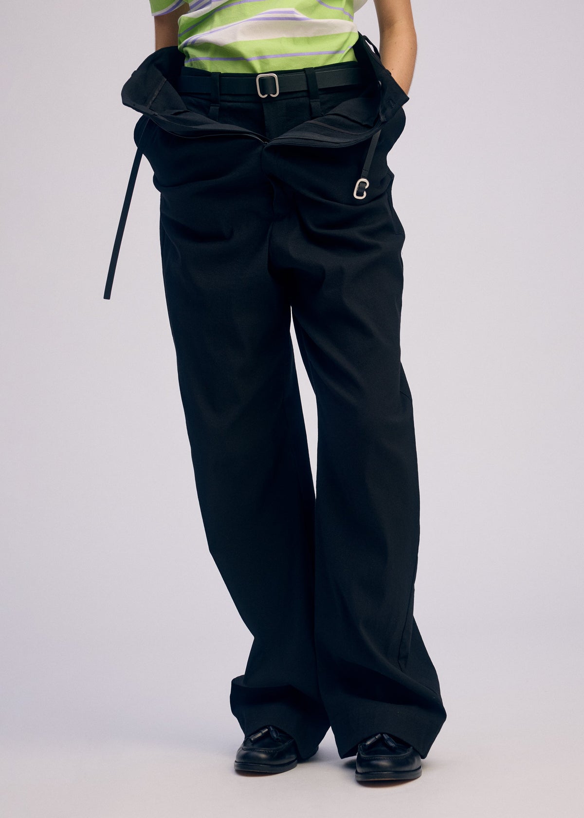 A person wears ISSEY MIYAKE GENERIC WEAR—oversized black, high-waisted pants with a belt, paired with a green and white striped top and black shoes. The loose trousers bunch at the waist and trail on the floor for a playful look. view 7 of 7