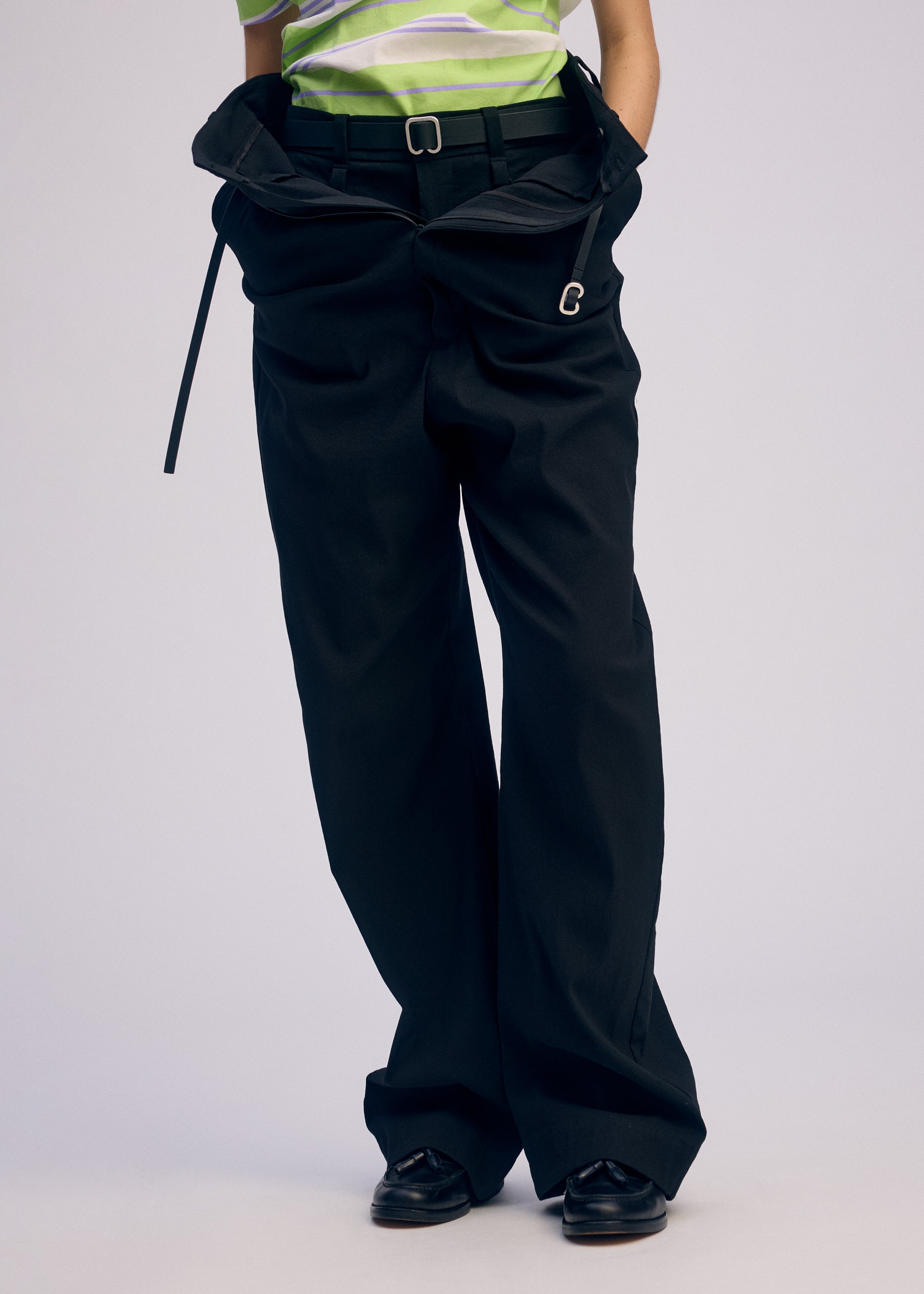 Zoomed view A person wears ISSEY MIYAKE GENERIC WEAR—oversized black, high-waisted pants with a belt, paired with a green and white striped top and black shoes. The loose trousers bunch at the waist and trail on the floor for a playful look.