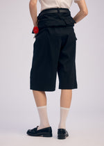 A model faces away wearing ISSEY MIYAKE GENERIC WEAR: high-waisted black knee-length shorts, a tucked-in white shirt, sheer white socks, black loafers, and a small red accessory on the shorts’ belt loop for a playful accent.