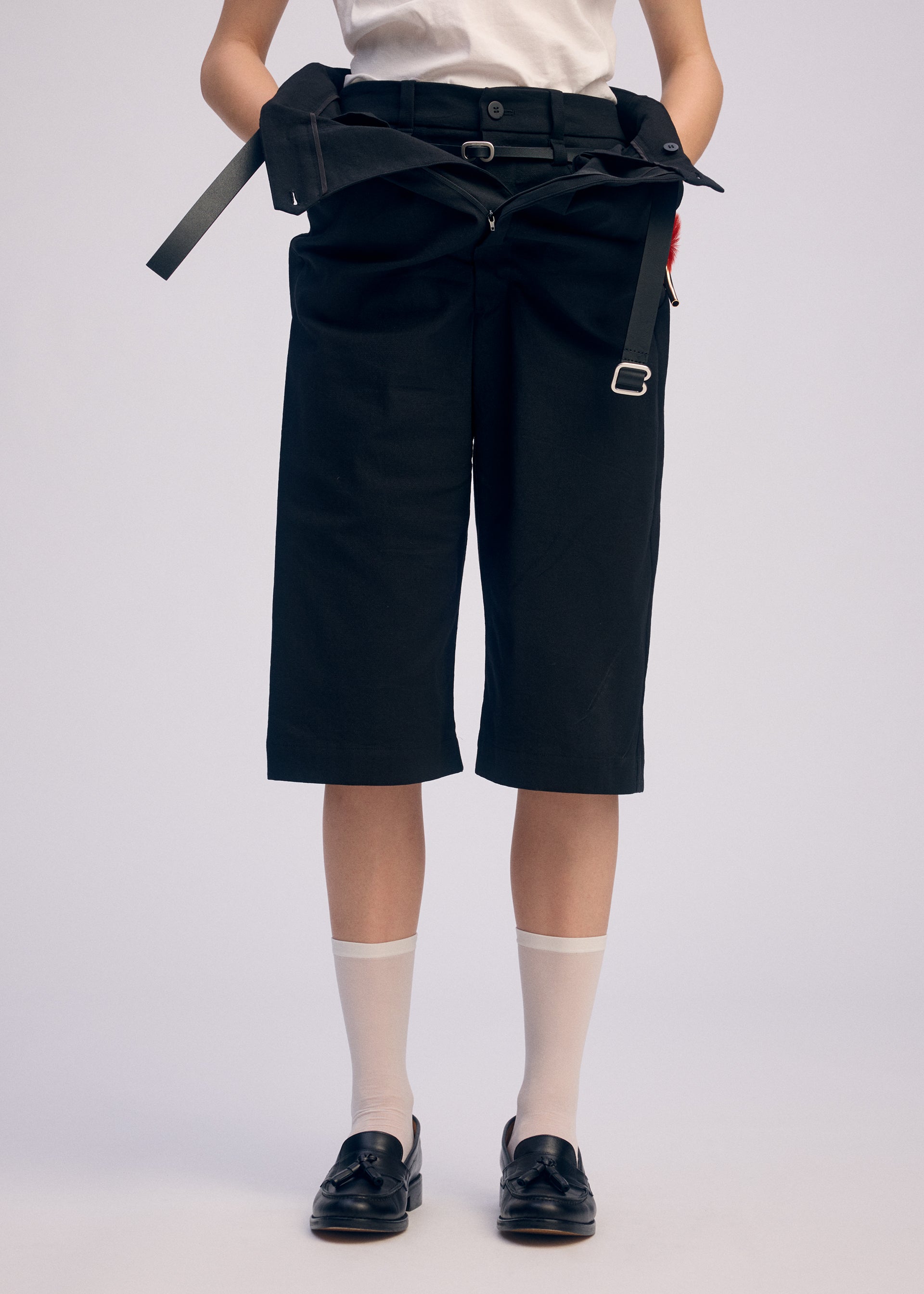Zoomed view Person dressed in ISSEY MIYAKE's GENERIC WEAR: black high-waisted shorts, white tucked-in shirt, black belt with silver buckle, white socks, and black loafers. Upper body mostly out of frame. A versatile styling choice.